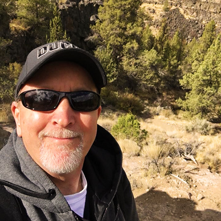 Jerry smiling outside wearing a jacket, sunglasses, and baseball hat surrounded by brush and evergreens.